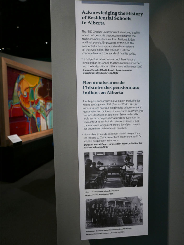 A view of one of the text panels at the residential schools exhibit is titled Acknowledging the History of Residential Schools in Alberta, noting the schools were part of “a policy of cultural genocide designed to dismantle the traditions and cultures of First Nations, Métis and Inuit people.” In the background is a partial view of Alex Janvier's large two-sided canvas <em>Blood Tears</em> (2001), relating to his experiences at Blue Quills Residential School. Photo: Tanya Harnett.