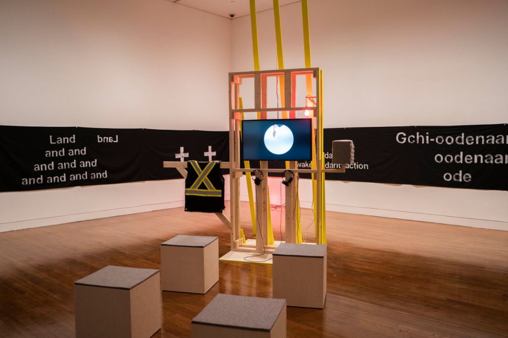 Maria Hupfield, <em>Electric Prop and Hum Freestyle Variations</em>, 2017–18. Wooden structure with hinged gate, 13 fluorescent-yellow wooden planks, felt objects, pink light fixture, “Land Acknowledgement banner” and video. Installation view of a performance at the Museum of Arts and Design, New York, 2017. Collaboration with Tusia Dabrowska. Courtesy the artist. © Maria Hupfield. Photo: Heidi Bohnenkamp, courtesy the Museum of Arts and Design, New York