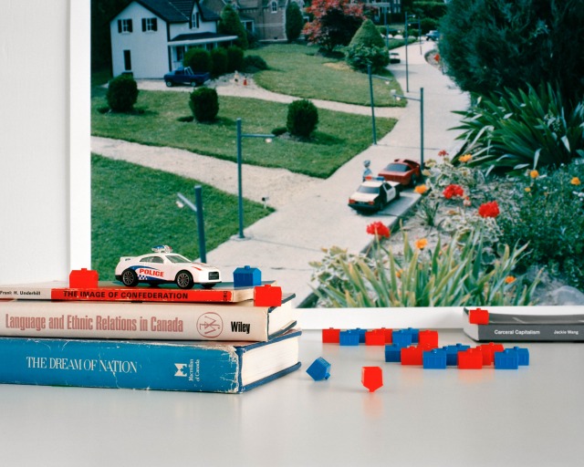 A toy police car rests on three books about nationhood; red and blue plastic houses sit in front of a photo of police in suburbia