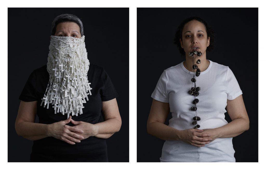 Two women, one with half her face wrapped in white rosaries, the other holding hair-wrapped stones in her teeth