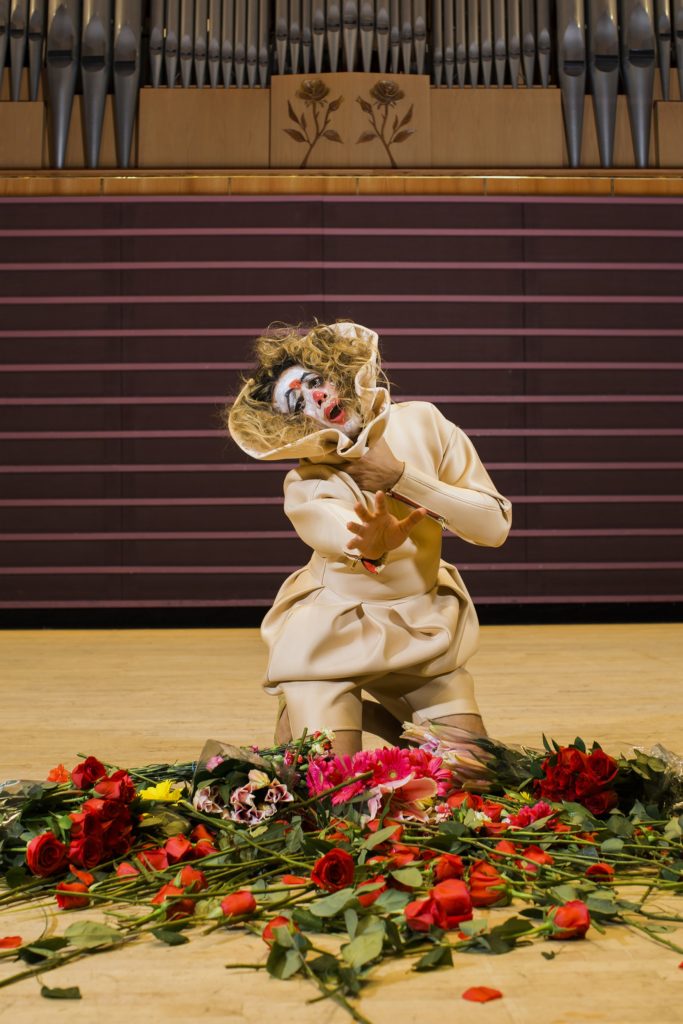 A woman in cream clothing and clown makeup kneels by a pile of roses onstage