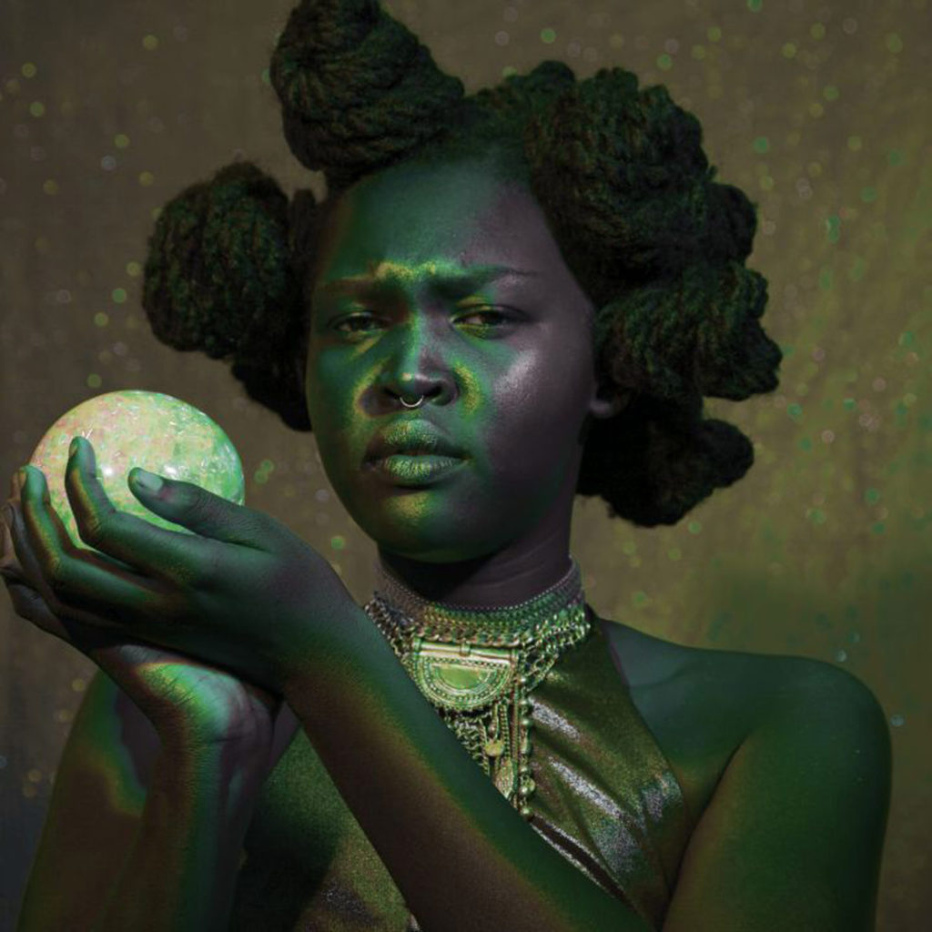 A Black femme person holds a pale orb and looks at the viewer