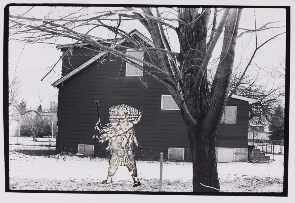 A multi-armed, multi-faced figure stands in front of a boarded-up house